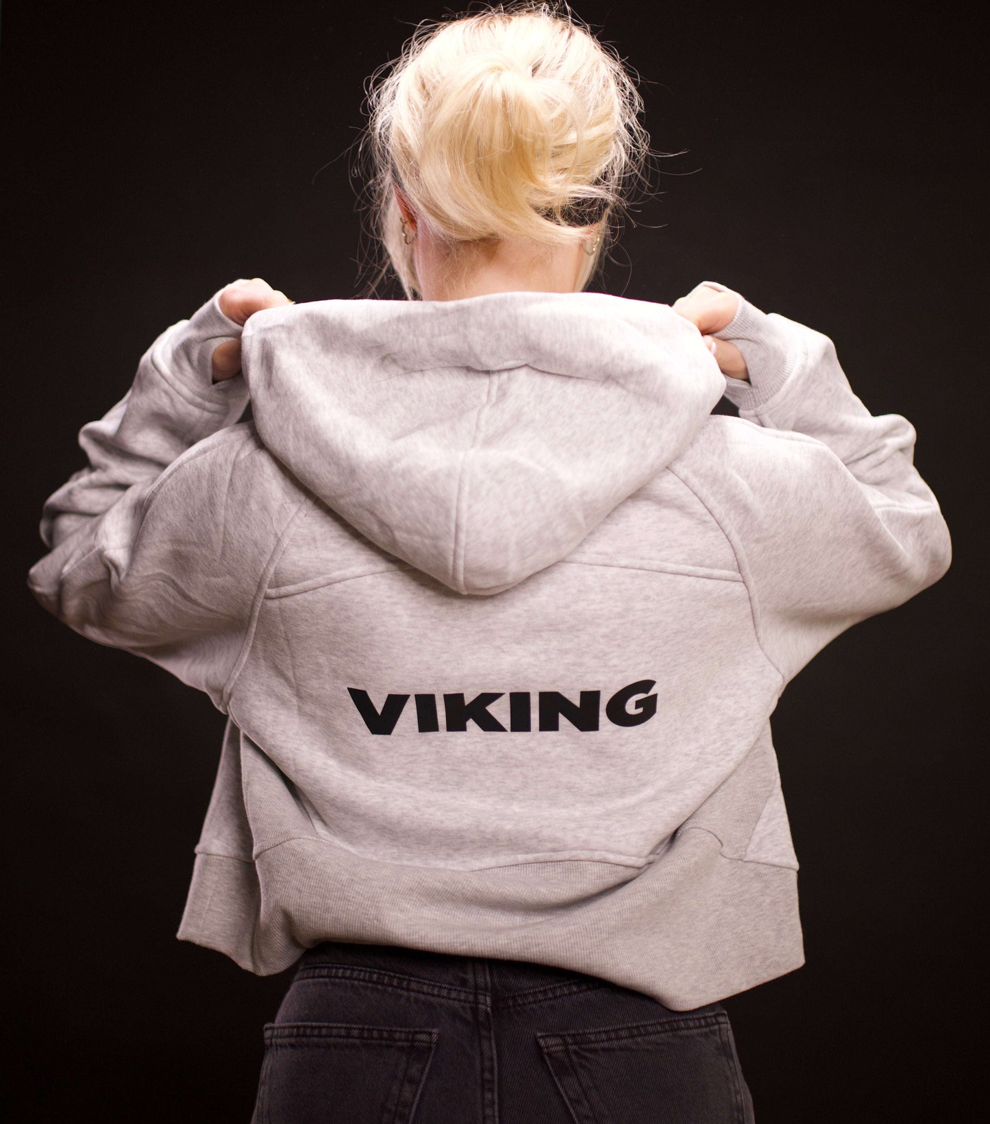 woman wearing light gray cropped Viking hoodie with hood up showing black Viking text on back against dark background
