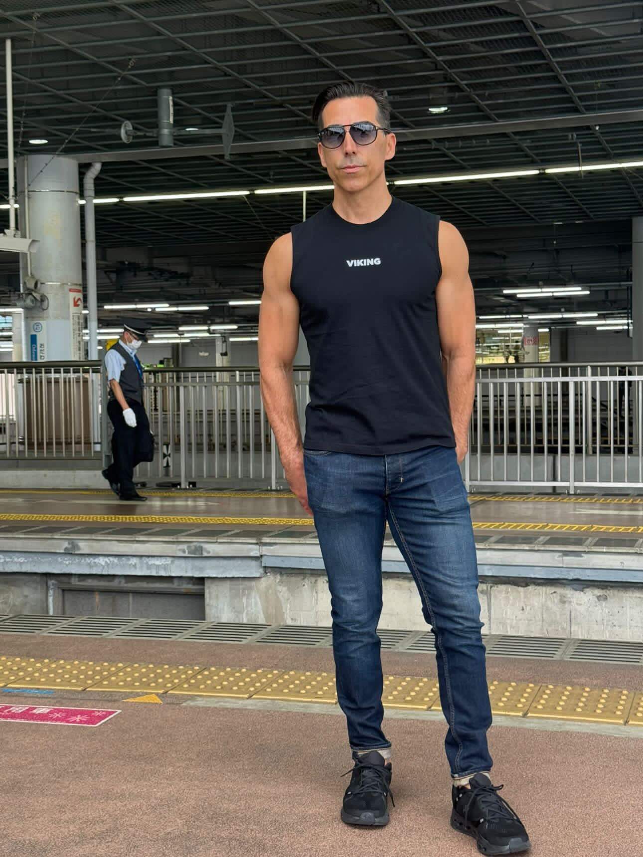Man wearing black Viking Tank Top with jeans and sunglasses at train station platform