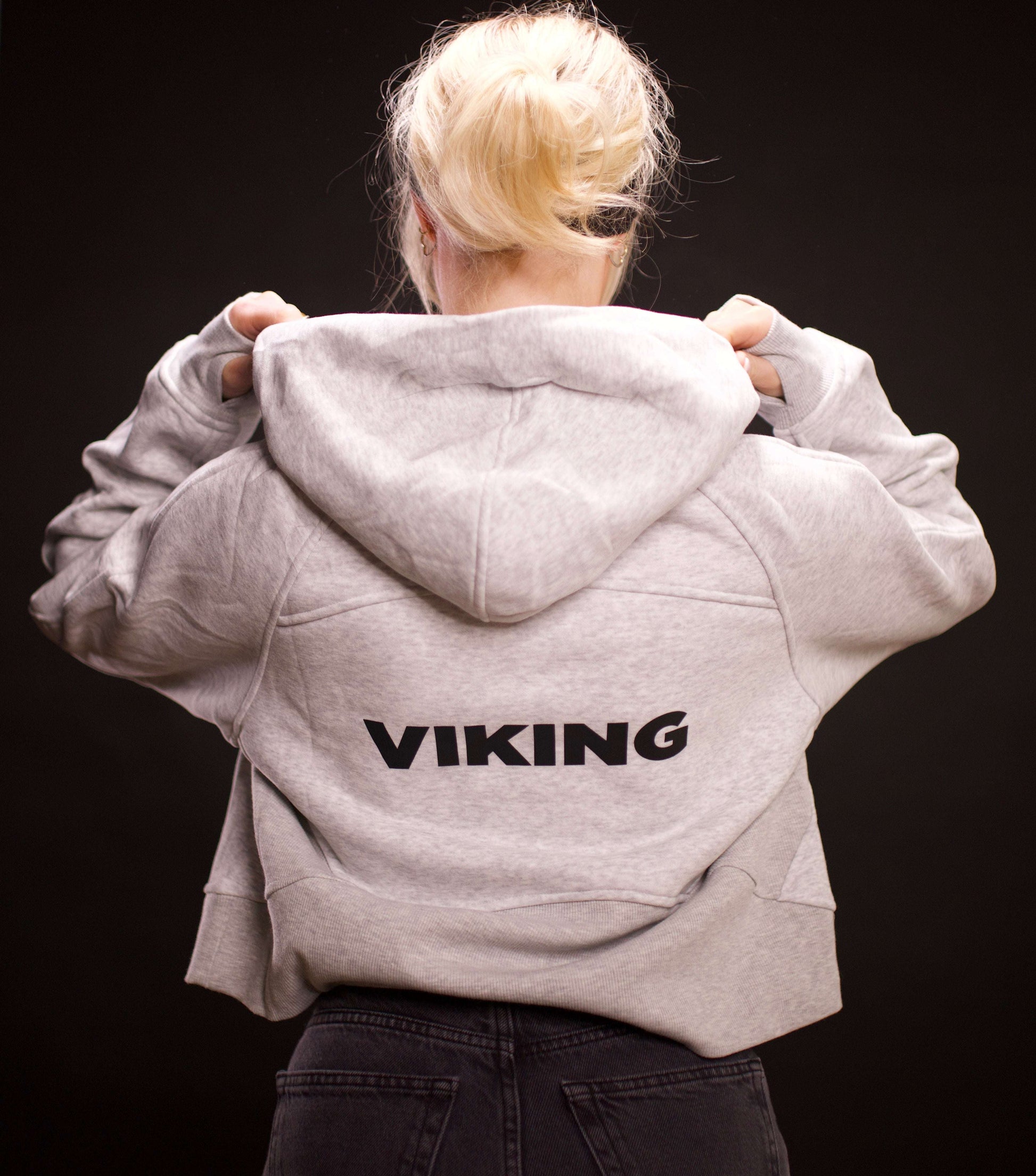 woman wearing light gray cropped Viking hoodie with hood up showing black Viking text on back against dark background